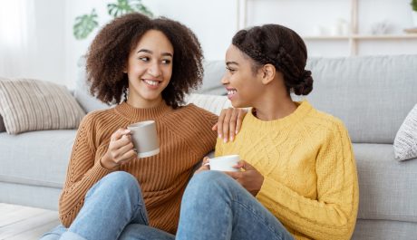 Females communication, gossip and modern date at home. Cheerful young african american women talk about relationships, discuss news, sit on floor near couch with cups of hot drink in living room