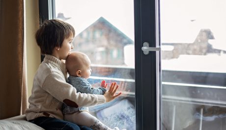 Preschool boy, holding his baby brother, sitting by the window in living room, looking at a snowy landscape outdoors